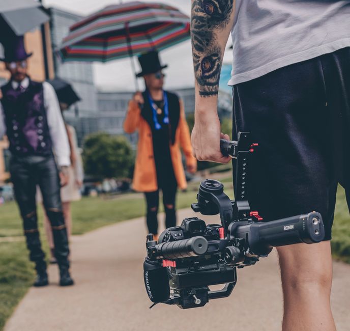 Pessoa segurando uma câmera de filmagem de alto nível em um ambiente ao ar livre, com duas outras pessoas ao fundo e uma pessoa com guarda-chuva colorido e chapéu.