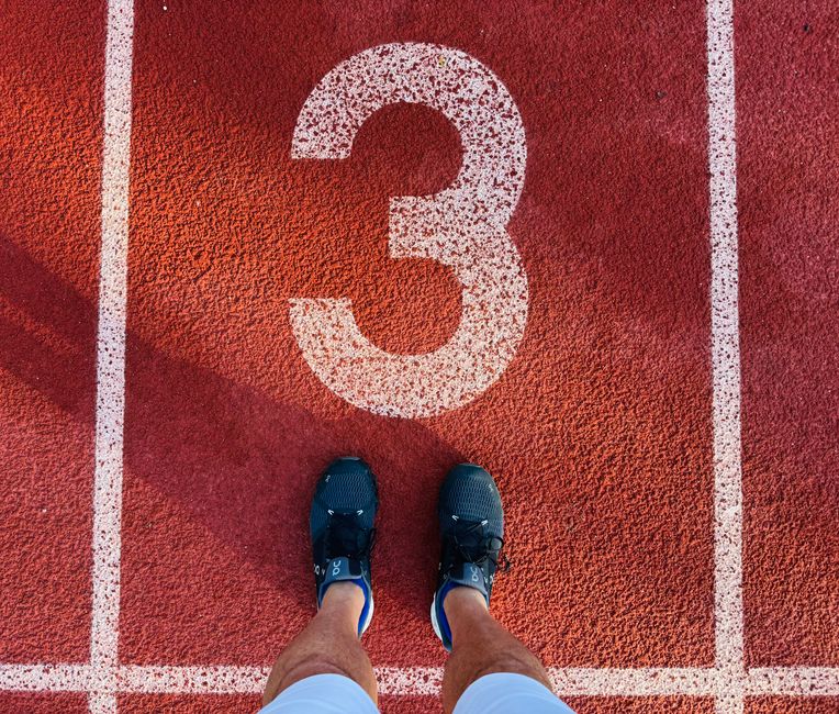 Imagem de uma pessoa de tênis em um local de corrida ou pista de atletismo, destacando o número 3 pintado no chão, simbolizando preparação ou chegada em uma competição.