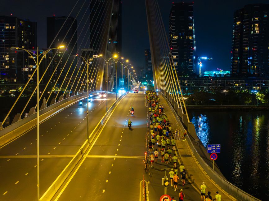 Imagem de uma ponte iluminada à noite com um grupo de corredores participando de uma maratona urbana na cidade.
