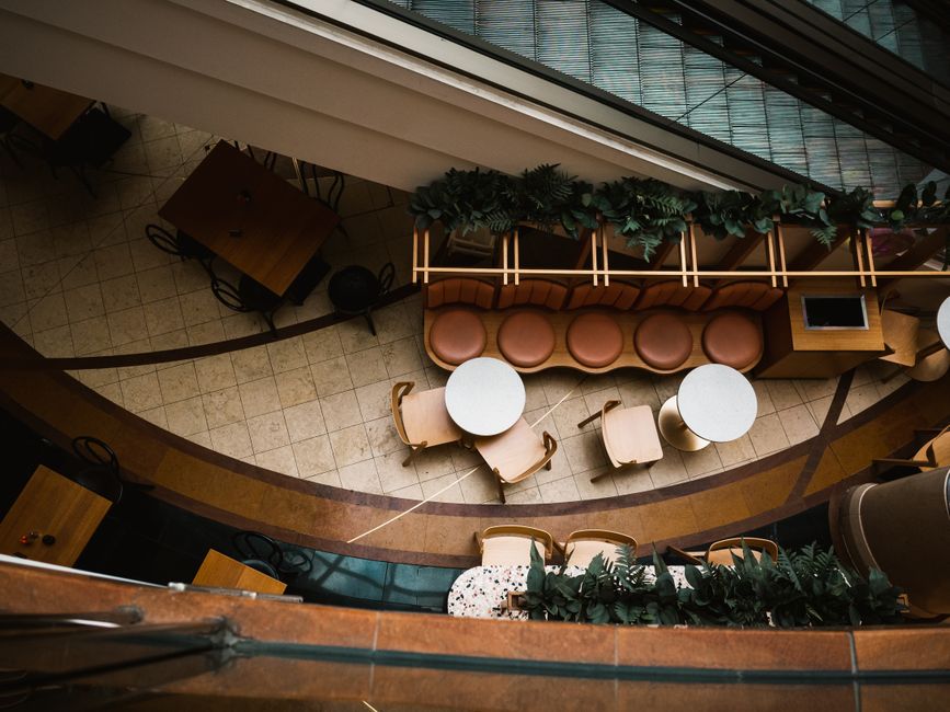 Imagem de um espaço de convivência em um ambiente corporativo, com mesas, cadeiras e uma área de descanso decorada com plantas, vista de cima para baixo.