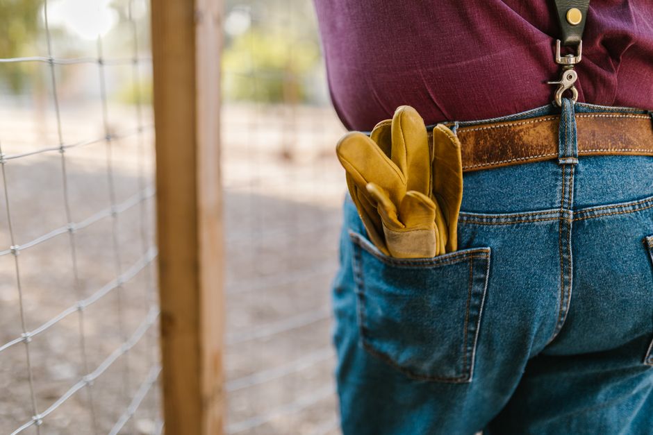 Homem segurando uma luva de trabalho na calça ao lado de uma cerca, em ambiente externo, representando trabalho na construção ou manutenção ao ar livre.