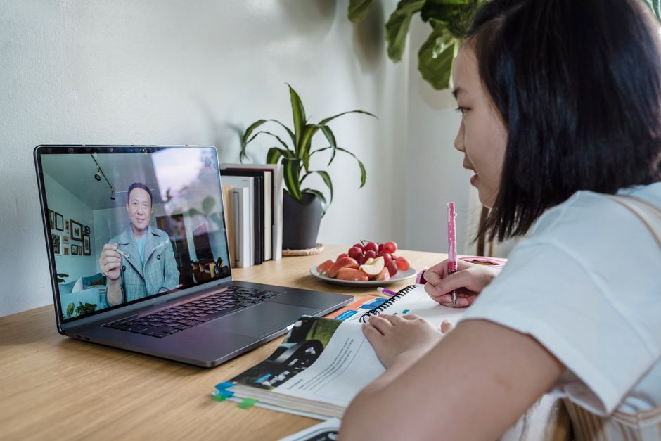 Pessoa assistindo a uma videochamada em um computador, enquanto estuda ou trabalha em um ambiente doméstico com plantas e livros ao fundo.