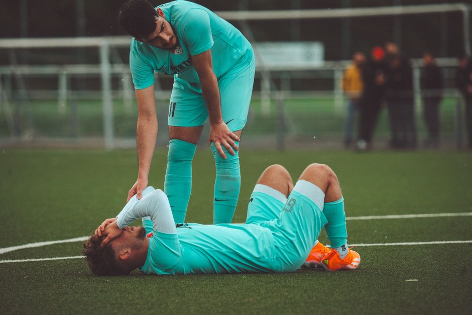 Jogador de futebol se recuperando de uma lesão enquanto um companheiro oferece ajuda no campo de jogo.