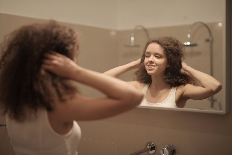 Jovem sorridente observando seu reflexo no espelho enquanto ajusta os cabelos no banheiro, transmitindo autoconfiança e autoestima.