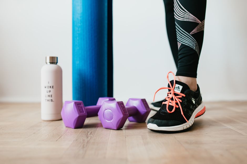Imagem de uma pessoa se preparando para a prática de exercícios físicos, com halteres, uma garrafa de água e um tapete de yoga ao fundo, enfatizando temas de saúde e bem-estar.