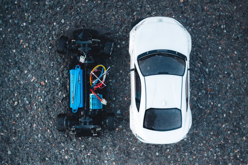 Imagem de um carro branco visto de cima ao lado de um veículo de controle remoto em terreno de asfalto, simbolizando tecnologia e inovação automotiva.
