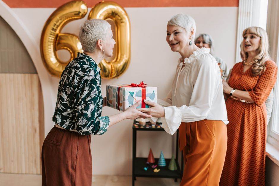 Mulher recebendo presente em celebração de aniversário entre amigas, com decoração festiva ao fundo.
