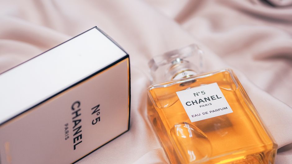 Imagem de um frasco de perfume Chanel No. 5 ao lado de uma caixa do mesmo produto, sobre tecido bege, transmitindo elegância e luxo.