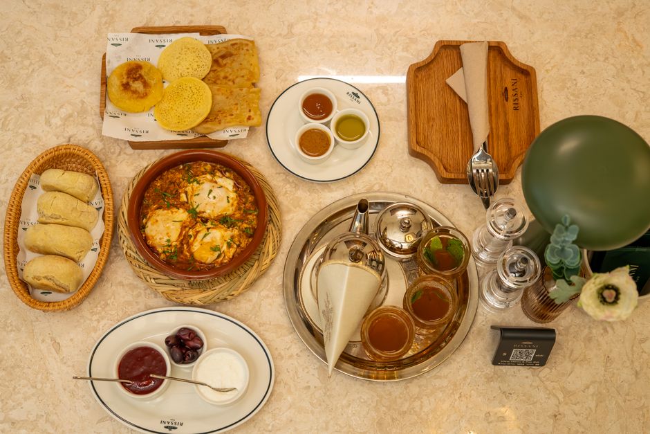 Mesa de café da manhã com variedades de pães, geleias, cafés e acompanhamentos, apresentando uma experiência gastronômica acolhedora.