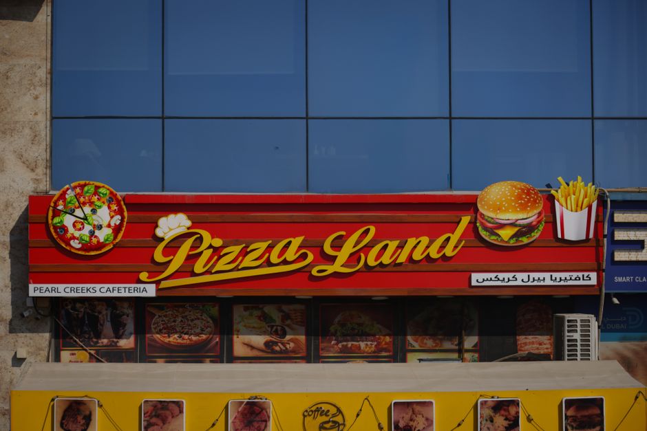 Imagem de uma fachada de restaurante de fast food com sinalização colorida e ilustrativa de pizza, hambúrguer, batatas e doces, em ambiente urbano.
