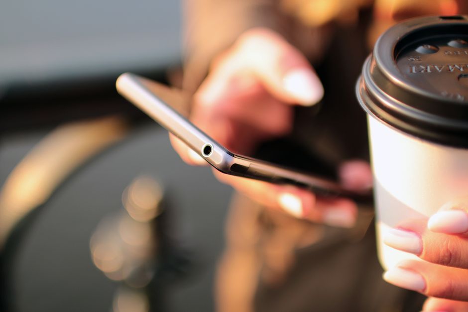 Pessoa usando smartphone e tomando café em ambiente ao ar livre, simbolizando produtividade e momentos de pausa na rotina.