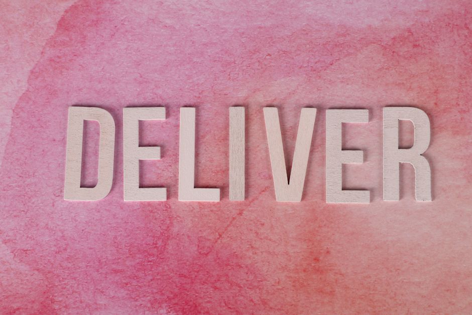 Imagem que destaca a palavra 'DELIVER' em blocos de madeira sobre um fundo rosado, simbolizando o conceito de entrega ou entrega de serviços.