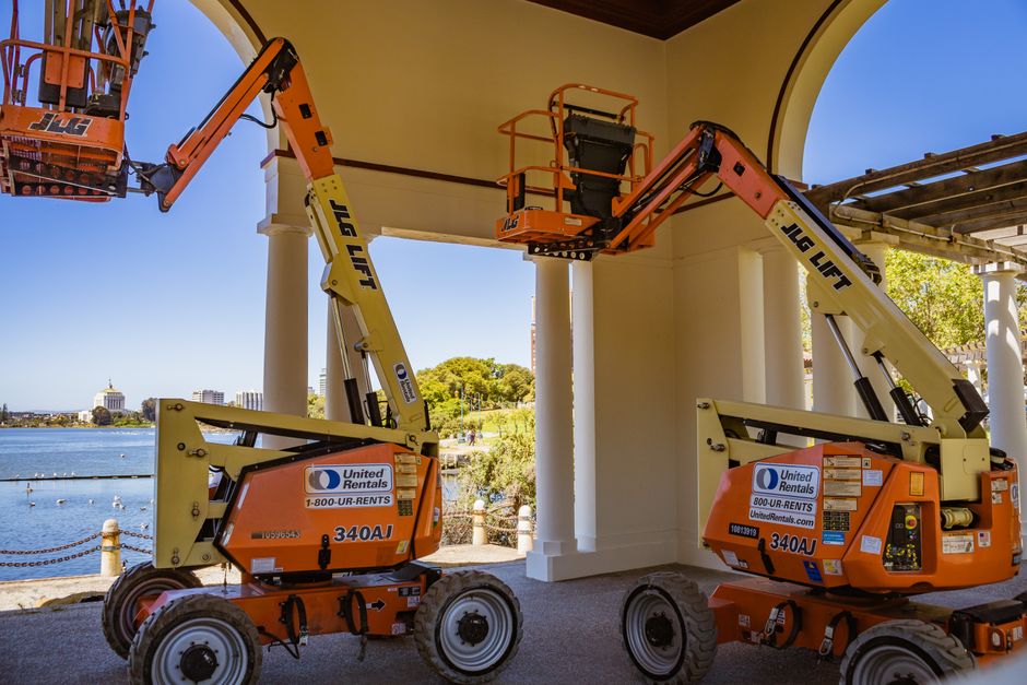 Duas plataformas elevatórias sendo usadas para realizar trabalhos de manutenção em uma estrutura sob um arco, com vista para um lago ao fundo e céu azul claro.