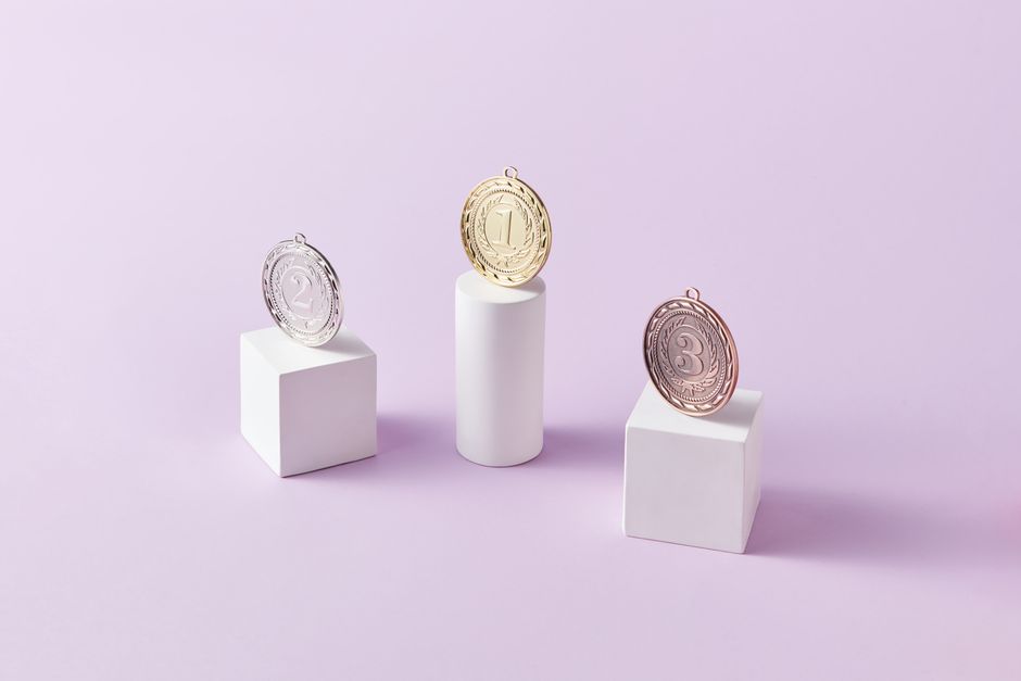 Medalhas de diferentes cores representando conquistas ou premiações, dispostas em posições de destaque sobre suportes brancos com fundo lilás suave.