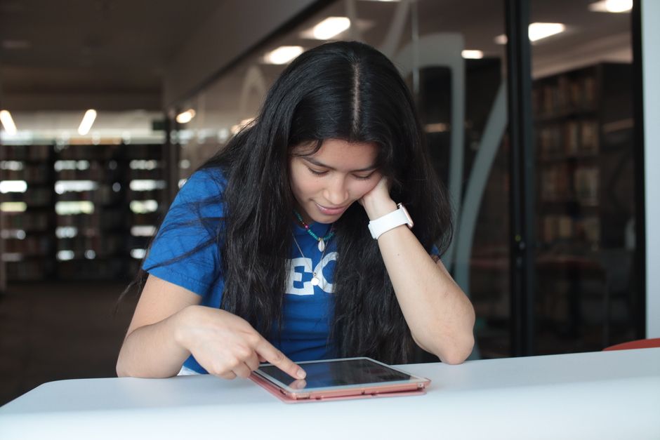 Jovem estudando ou pesquisando em biblioteca moderna, utilizando um tablet portátil com foco na busca por conhecimento ou estudo digital.