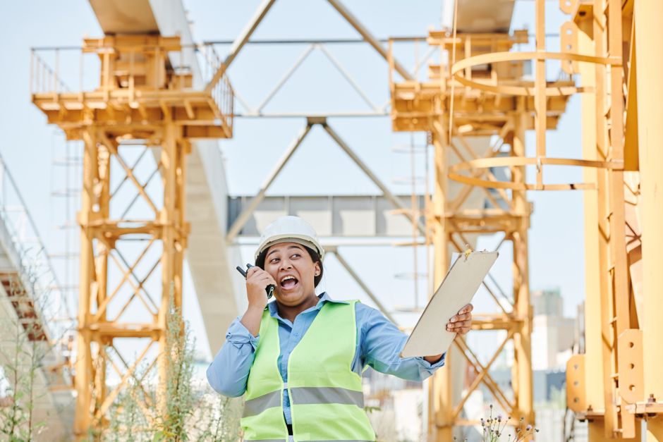 Engenheira de construção falando ao telefone e segurando uma prancheta em um canteiro de obras ao ar livre, durante uma inspeção de segurança ou supervisão de projeto.