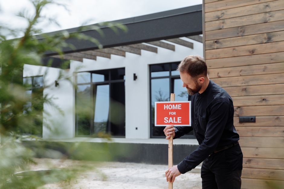 Homem segurando placa de anúncio de venda de imóvel em frente a uma casa moderna, destacando o mercado imobiliário.