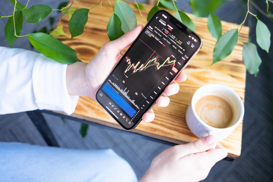 Pessoa analisando gráficos de ações em smartphone durante o dia com uma xícara de café ao lado, em um ambiente com plantas e madeira, simbolizando finanças e investimentos.