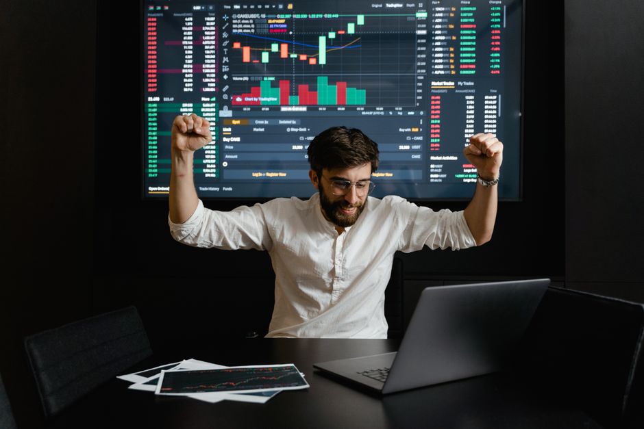 Homem celebrando vitória no mercado financeiro enquanto analisa gráficos de ações em um ambiente de trabalho moderno.