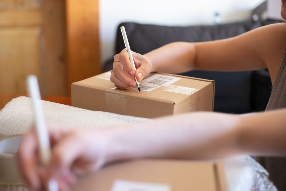 Pessoa preenchendo etiqueta de envio em uma caixa de entrega, simbolizando logística e operações de comércio eletrônico.