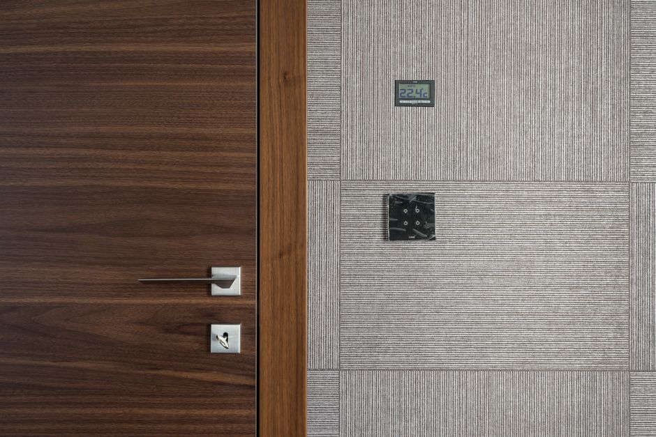 Imagem de uma porta de madeira ao lado de uma parede com um painel de controle digital e um relógio analógico, transmitindo um ambiente corporativo moderno.
