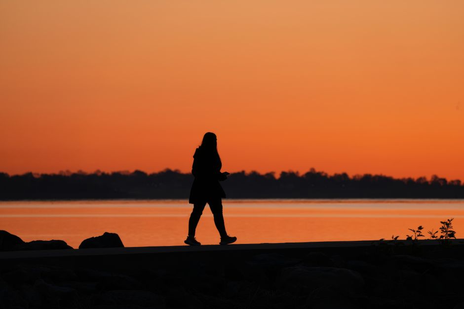 Silhueta de uma pessoa caminhando ao pôr do sol à beira de um rio, com o céu em tons de laranja e rosa, transmitindo calma e reflexão.