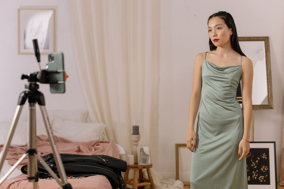 Mulher posando para gravação de vídeo em ambiente de quarto, com uma câmera em foco à esquerda.