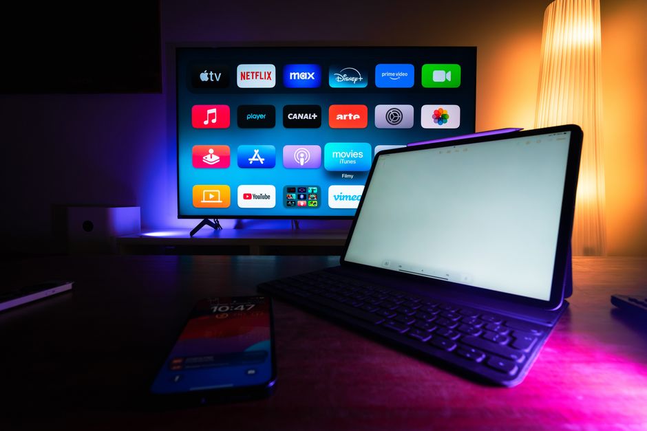 Imagem de um espaço de entretenimento com uma televisão moderna exibindo aplicativos de streaming e um dispositivo móvel com teclado auxiliar, iluminado por luzes ambientas quentes e frias.