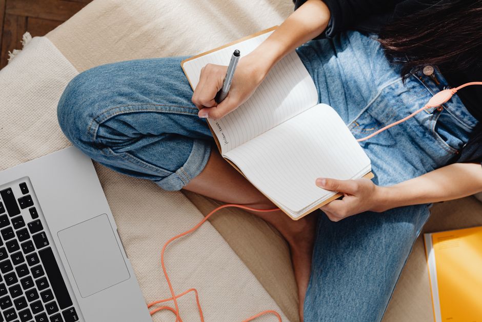 Pessoa anotando ideias ou planejamento em um caderno, com laptop ao lado, em um ambiente confortável e informal.