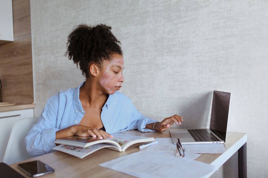 Mulher estudando e realizando pesquisa online em um ambiente de casa, combinando leitura e uso de computador.
