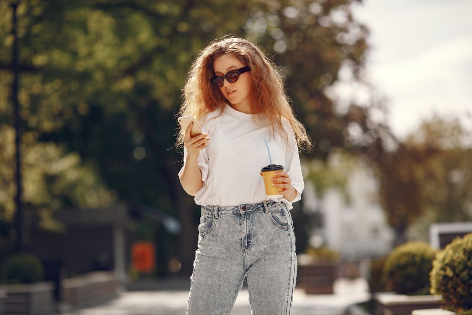 Mulher jovem usando óculos escuros e camiseta branca, caminhando ao ar livre enquanto utiliza o celular e segura uma bebida, em um ambiente urbano e ensolarado.