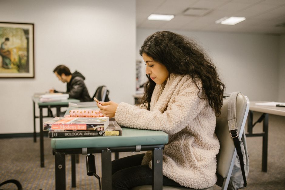 Mulher estudando ou lendo em uma sala de aula ou biblioteca, focada em seus estudos, com livros e materiais escolares na mesa.