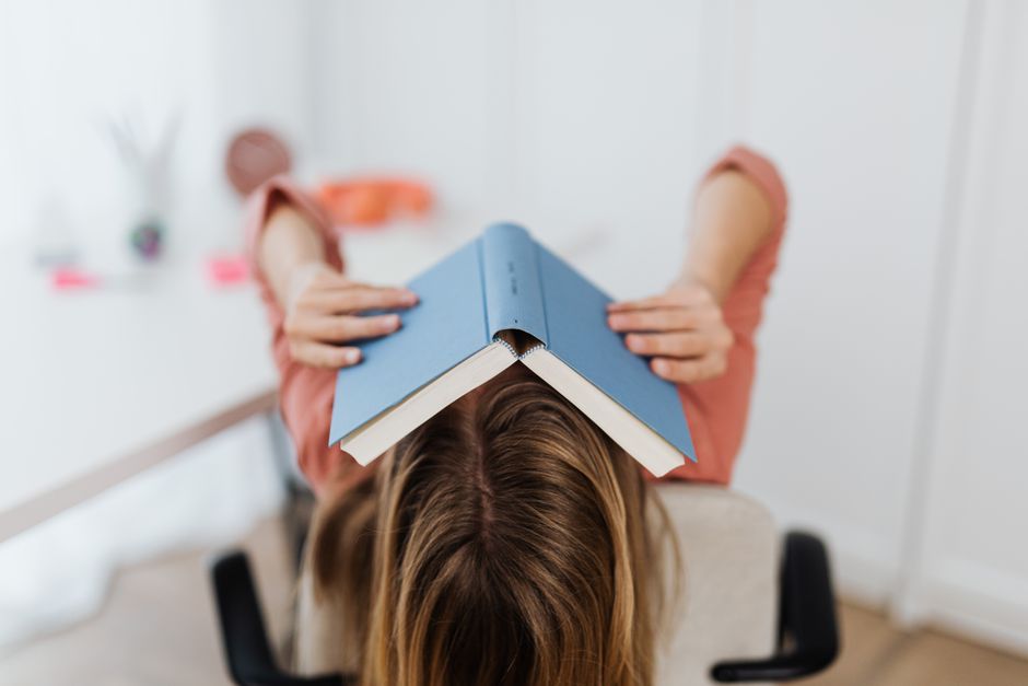Pessoa deitada com cabeça sobre uma mesa, usando um livro como uma touca, representando foco na leitura ou estudo intenso.