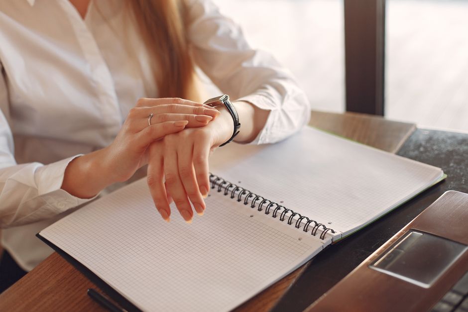 Mulher verificando o horário em um relógio enquanto faz anotações em um caderno, simbolizando gerenciamento de tempo e produtividade no ambiente de trabalho.