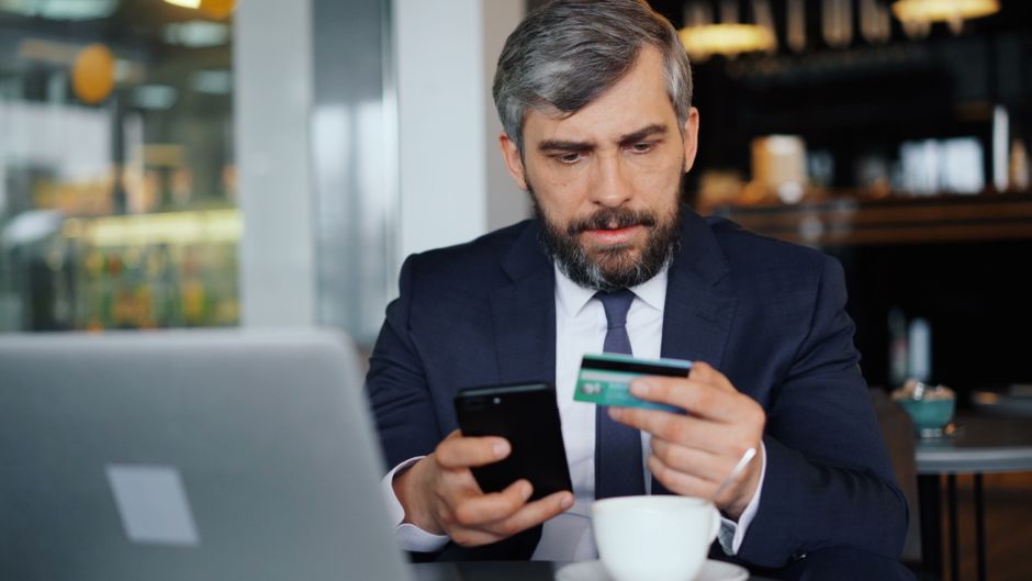 Homem de terno realizando uma compra online com cartão de crédito em um ambiente de café ou coworking, simbolizando assuntos relacionados a finanças, comércio digital e segurança nas transações.
