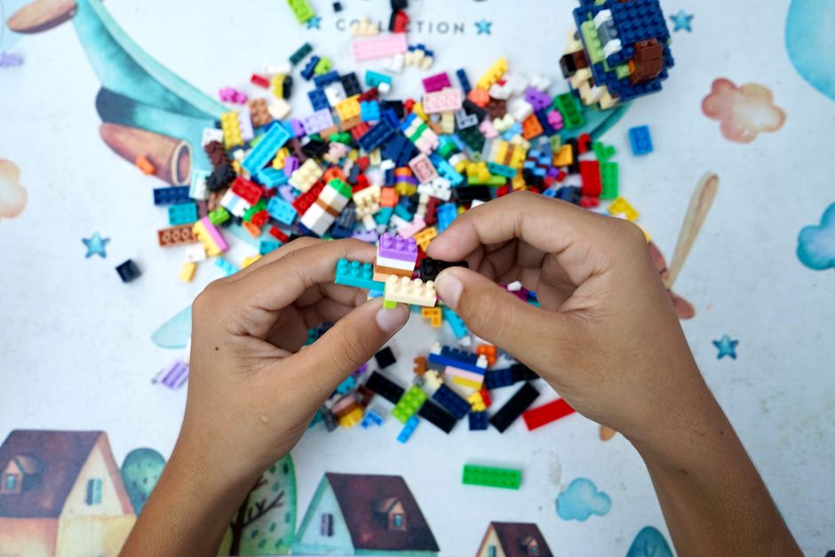 Mãos montando uma peça de lego colorida sobre uma mesa com peças espalhadas, simbolizando criatividade e desenvolvimento infantil.