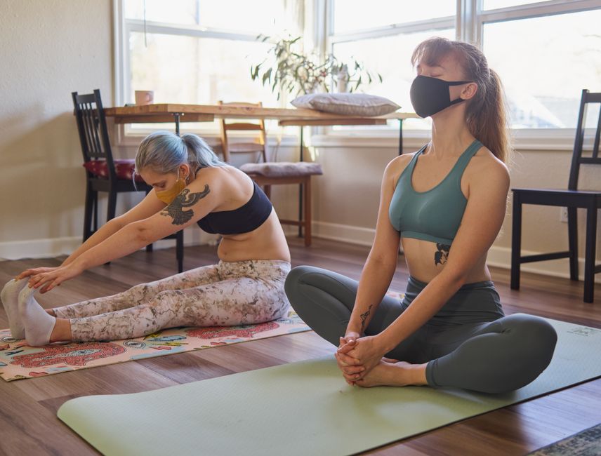 Duas mulheres praticando yoga em casa, com uma delas fazendo alongamento e a outra meditando, usando máscara facial.