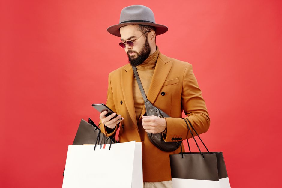 Homem elegante usando roupas modernas e acessórios com várias sacolas de compras, explorando o uso de tecnologia em compras e moda.