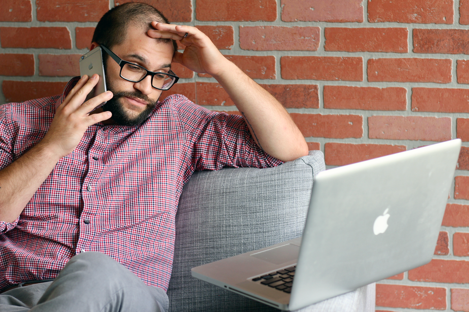 Homem preocupado trabalhando com computador portátil e telefone, ilustrando estresse ou desafio relacionado à tecnologia ou trabalho remoto.
