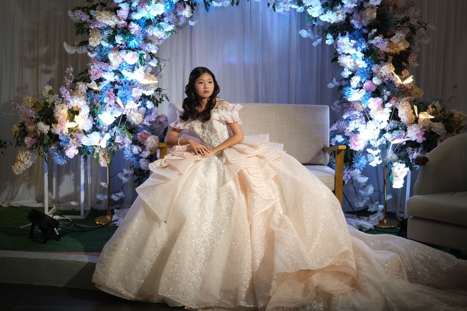 Menina em vestido de debutante sentada em um cenário decorado com flores para comemoração especial.