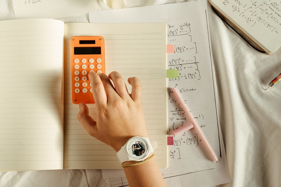 Pessoa estudando matemática ou cálculos avançados com uso de calculadora, caderno e livros de apoio.