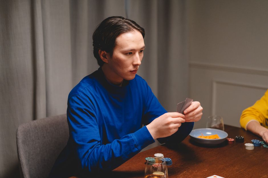 Jovem concentrado jogando pôquer em uma mesa de ambiente interno, com fichas e cartas na mão.