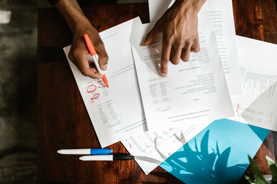 Pessoa revisando e marcando documentos financeiros ou de negócios com atenção aos detalhes, em um ambiente de trabalho organizado.