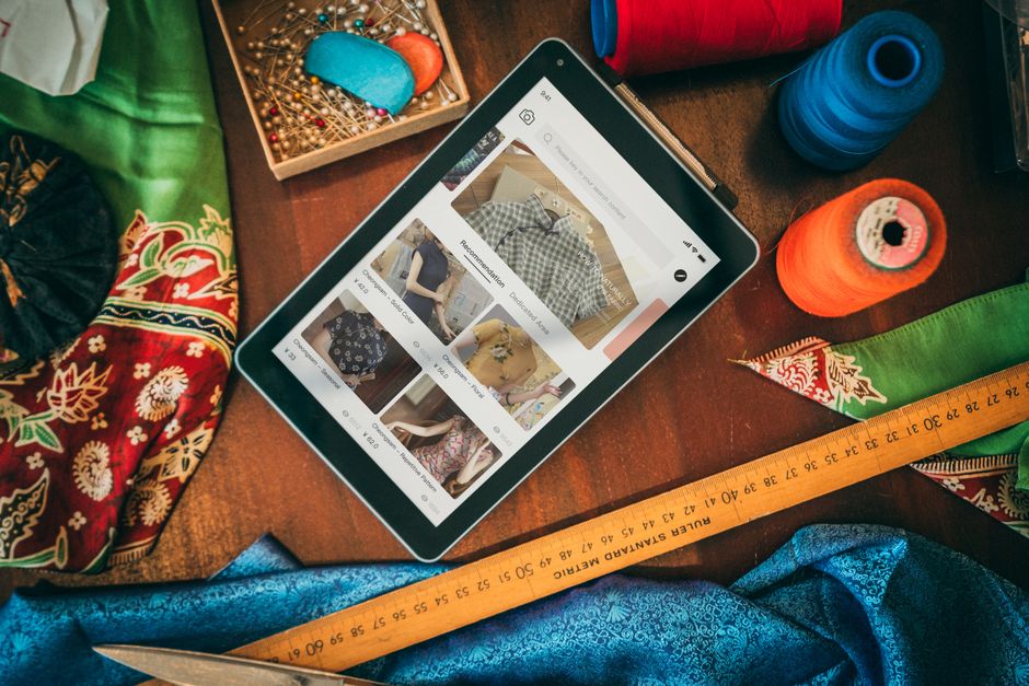 Imagem de uma mesa de costura com destaque para uma tela de tablet exibindo recomendações de roupas, rodeada por materiais como linhas, tecidos e agulhas, sugerindo produção e moda familiar.