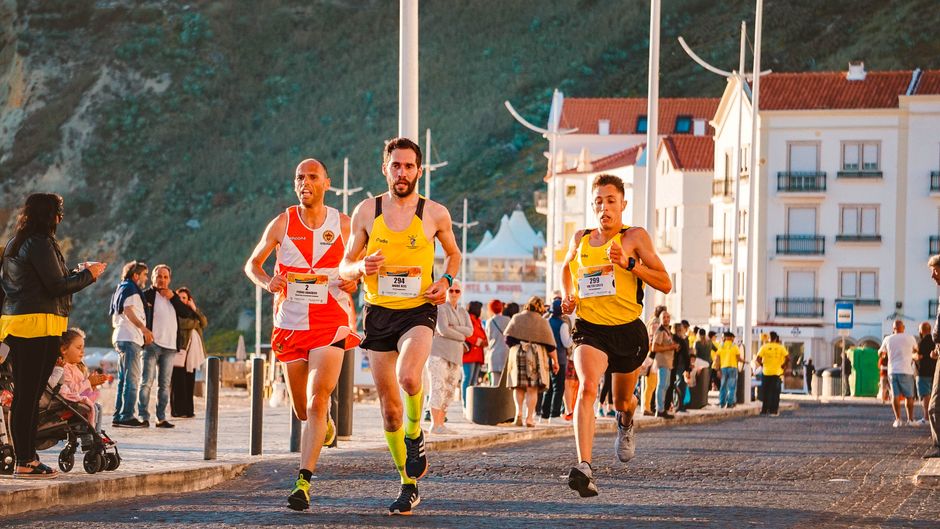 Três corredores participando de uma corrida de rua em um ambiente urbano ao pôr do sol, com espectadores assistindo ao evento.