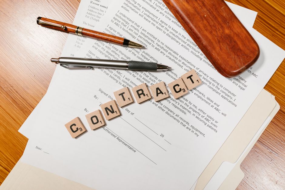 Documento de contratação com blocos de letras formando a palavra 'CONTRACT' sobre uma mesa de trabalho com canetas e uma caixa de madeira ao fundo.