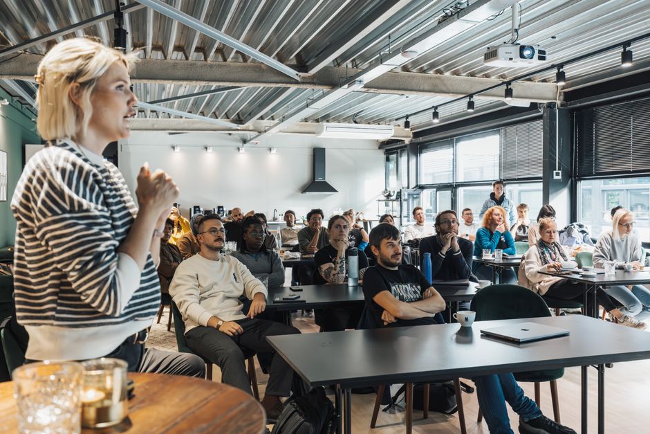 Mulher apresentando palestra para uma audiência diversificada em um ambiente moderno de coworking ou espaço de eventos.