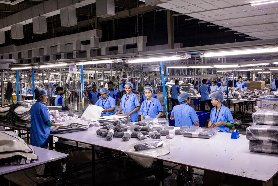 Imagem de uma fábrica de manufatura com trabalhadores em uniformes azuis, envolvidos na produção de itens têxteis ou estofados, destacando o ambiente de produção em grande escala.