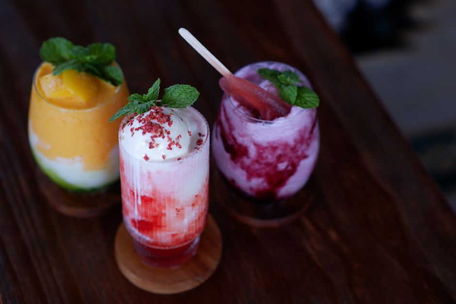 Três drinques coloridos decorados com hortelã sobre uma mesa de madeira, ideais para um momento de lazer ou celebração.