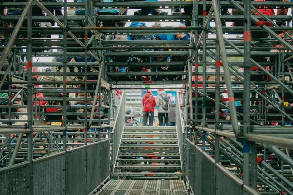 Imagem de uma estrutura de escadas e andaimes metálicos, com pessoas caminhando ao fundo, sugerindo uma área de construção ou evento ao ar livre.